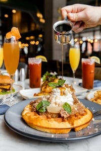 Maple syurp being poured by a server- just the hand is in the shot- on a chicken and waffles dish. With bloody marys and mimosas in the background